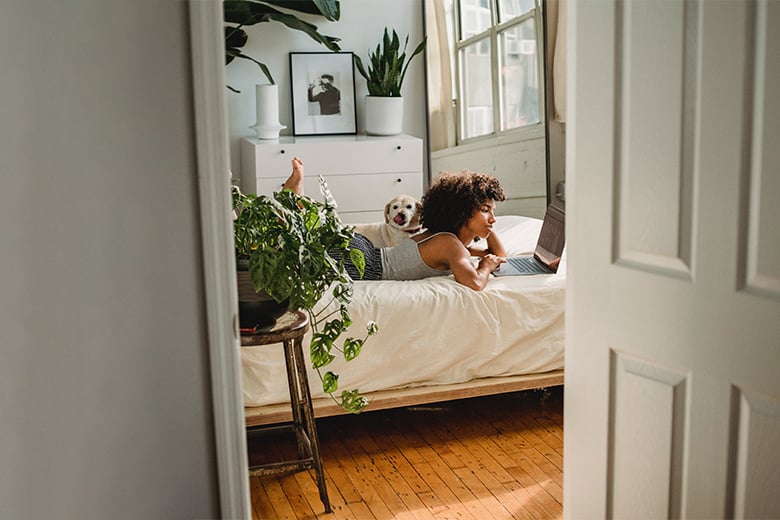 Kvinna ligger på en säng med sin hund och studerar onlinekurser på en dator