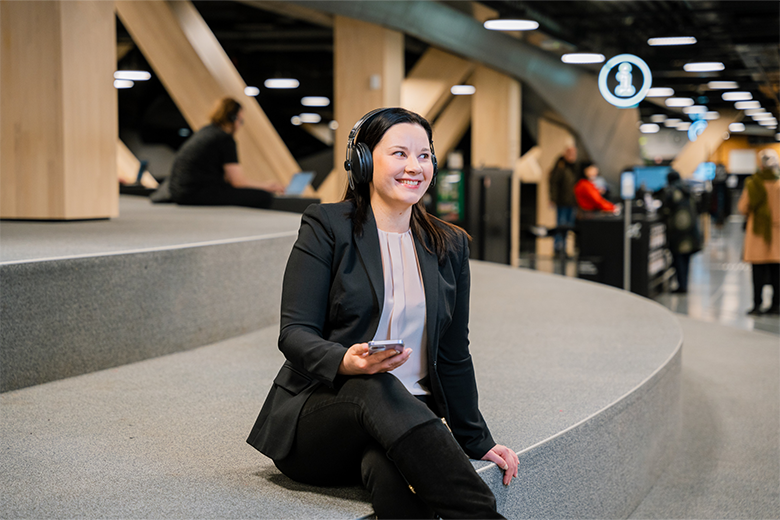 Kvinna sitter på en bänk i ett bibliotek och lyssnar leende på en podcast från sin mobiltelefon i ett par hörlurar