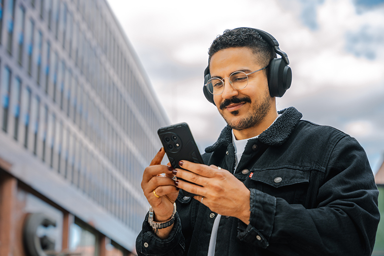 Man bär hörlurar och tittar leende ner i en mobiltelefon som han håller upp