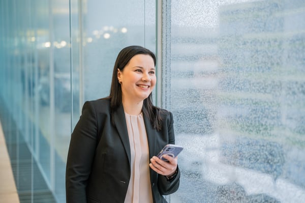 kvinna i rosa blus och kostym med mobiltelefon som ser glad ut när hon tittar ut genom glas